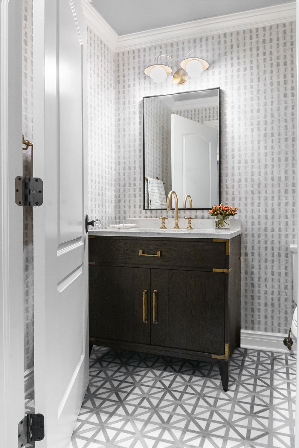 Small bathroom with dark wood vanity, gold accents, patterned wallpaper, and geometric floor tile.