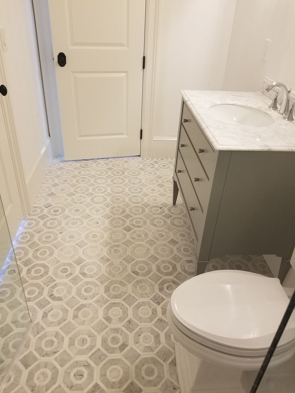 Bathroom with gray vanity, white toilet, and patterned tile floor. A white door is in the background.