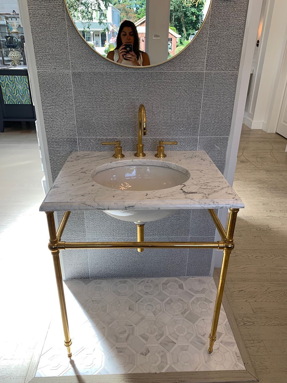 Gold-legged sink with white marble countertop and gold faucet, in front of a gray tiled wall and round mirror.