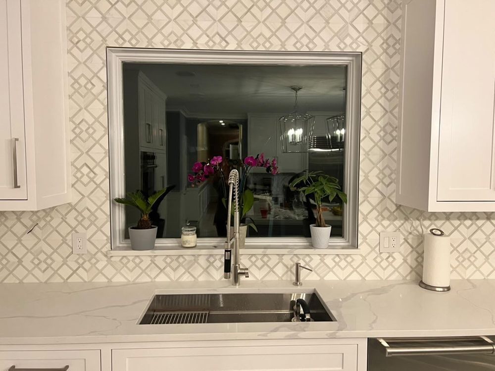 Kitchen sink with window above, backsplash of patterned tiles, white cabinets, and plants.