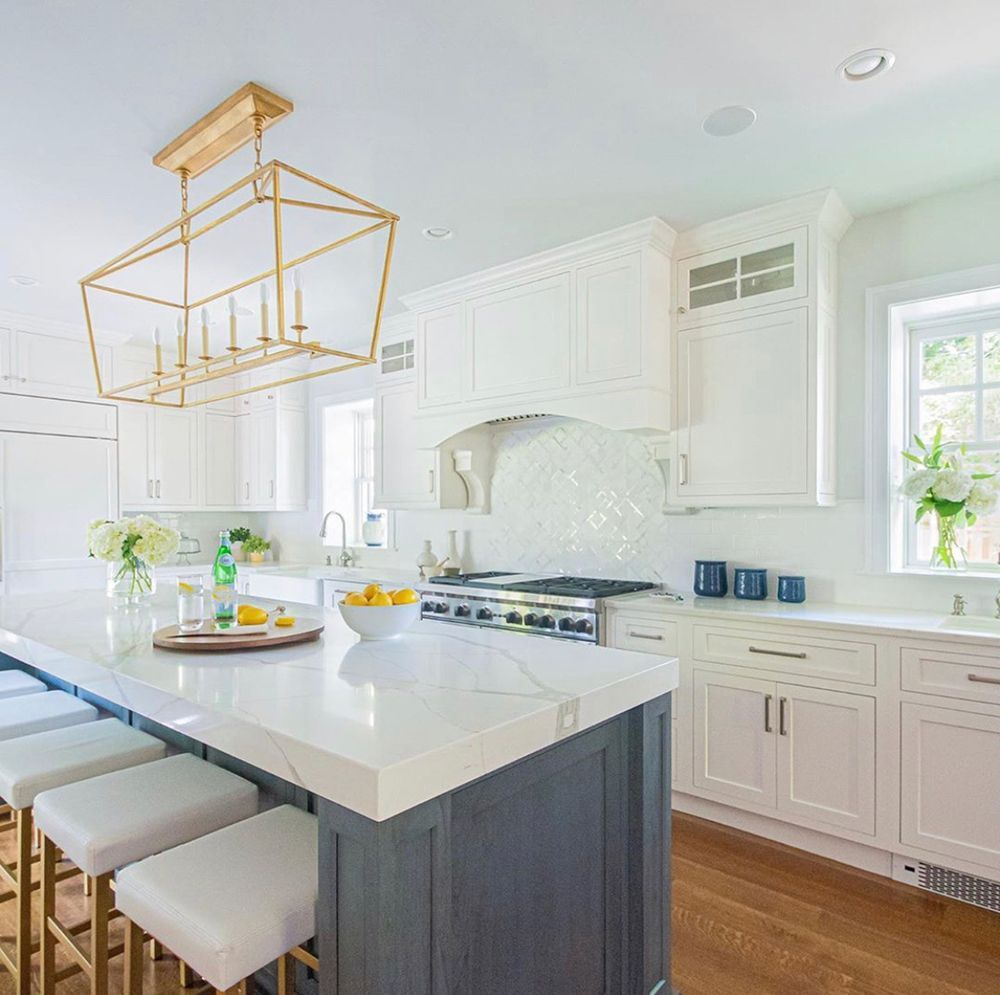 Modern white kitchen with a large island, brass chandelier, and wood floor.