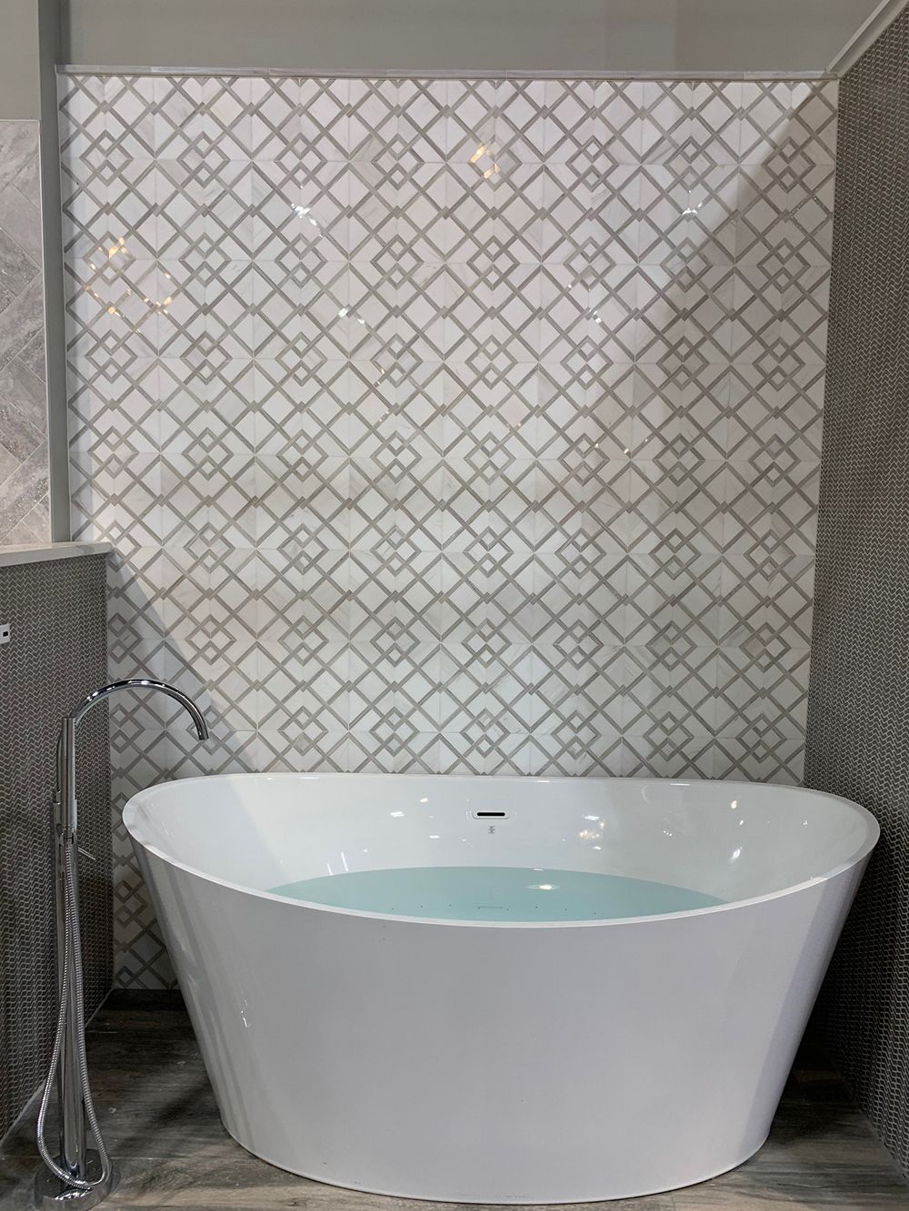 Freestanding white bathtub with water in front of a patterned tile wall.