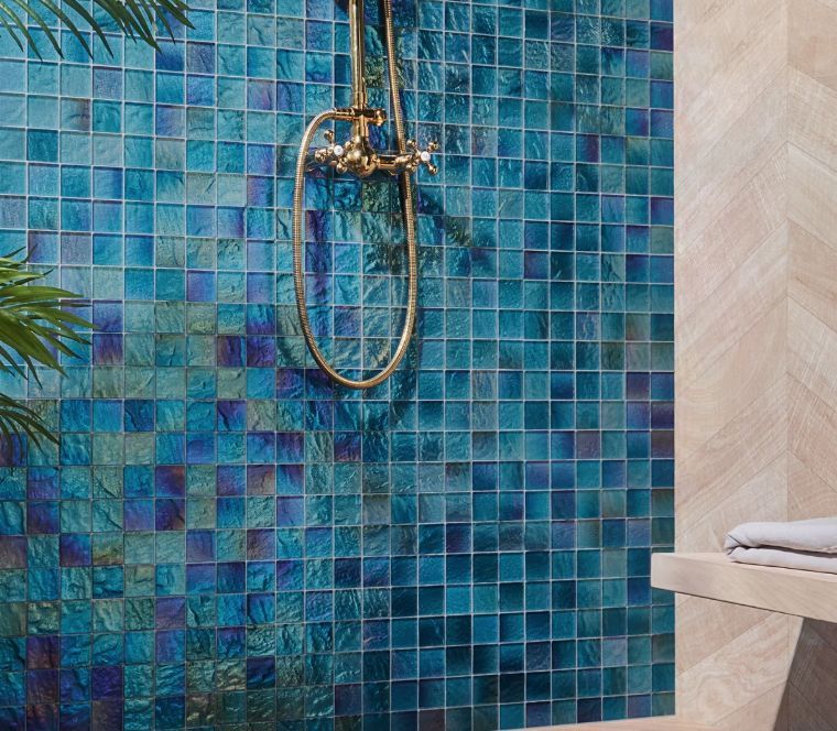 Blue iridescent square tiles on a shower wall with gold shower fixtures and a wooden accent wall.