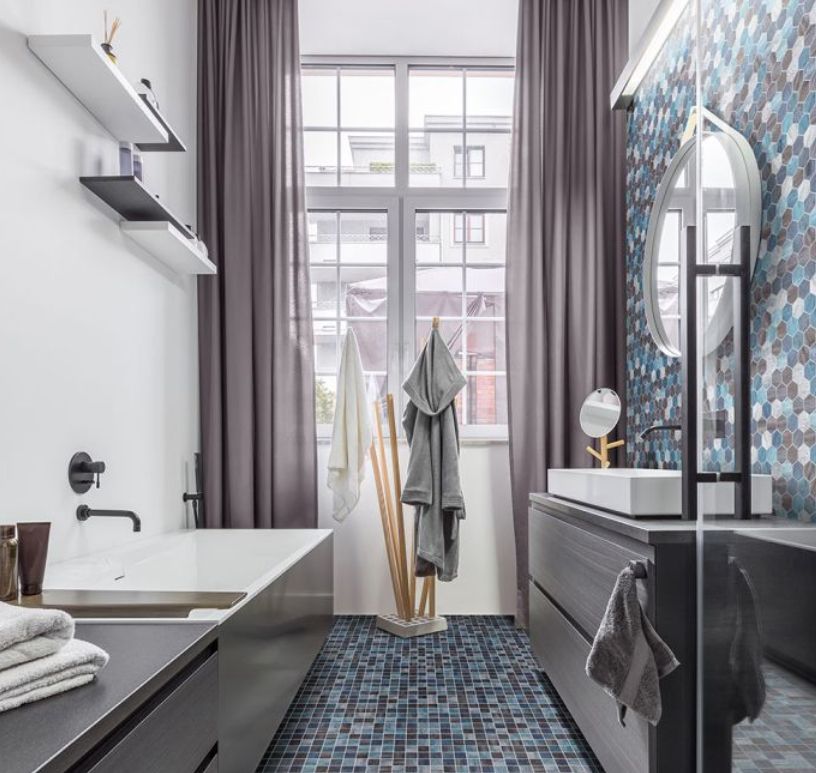 Modern bathroom with gray cabinets, tub, and mosaic tile flooring; window with purple curtains.