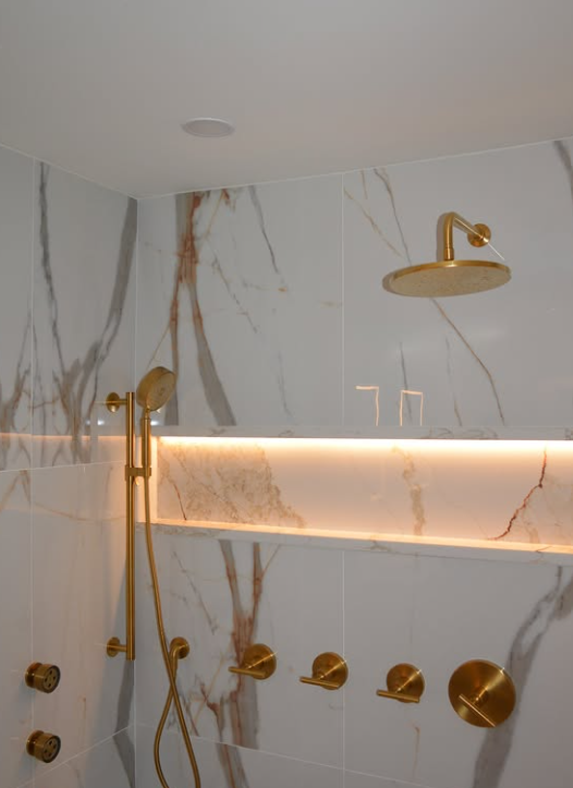Gold shower fixtures in a white marble-tiled shower, with a backlit shelf.