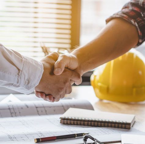Two people shaking hands over blueprints, a notepad, and a yellow hard hat.
