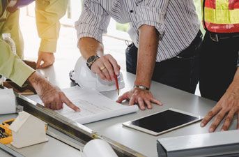 Construction workers review blueprints, pointing and discussing at a table with a tablet and hardhat.