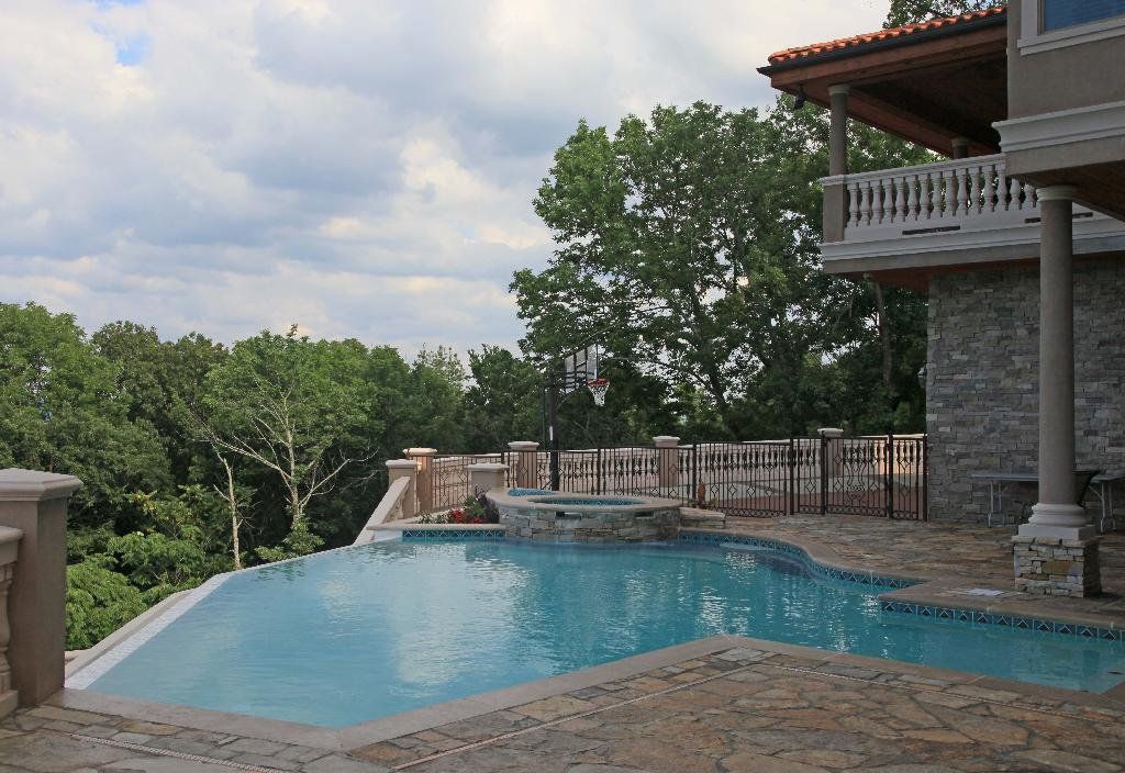 Pool with spa overlooking trees; stone patio, ornate house, cloudy sky.