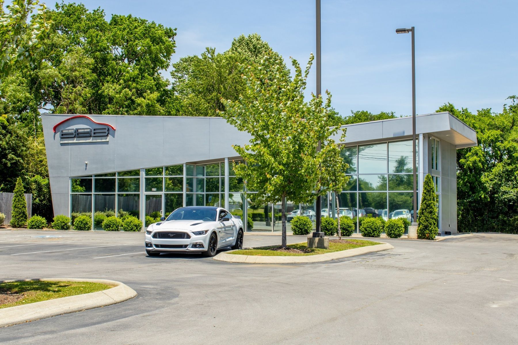 White Ford Mustang parked in front of a modern car dealership with large windows on a sunny day.