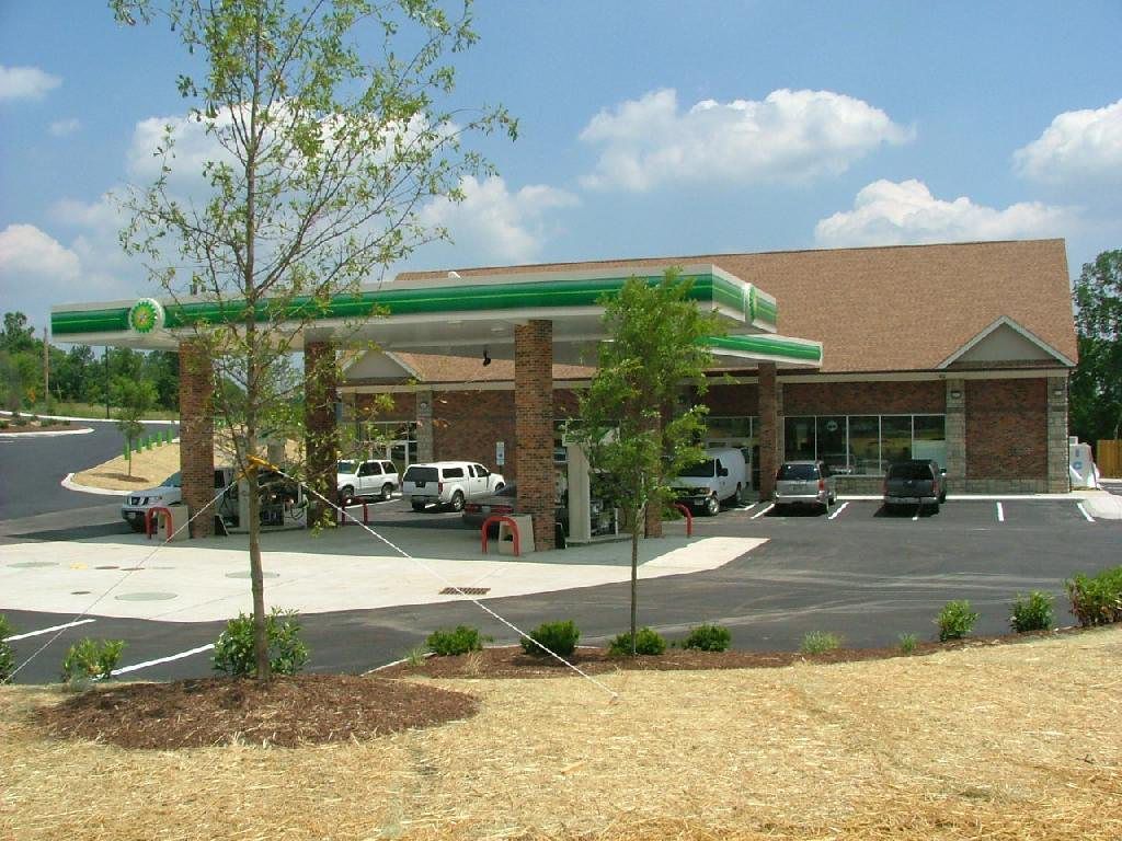 BP gas station with canopy, brick building, fuel pumps, vehicles, and trees under a blue sky.