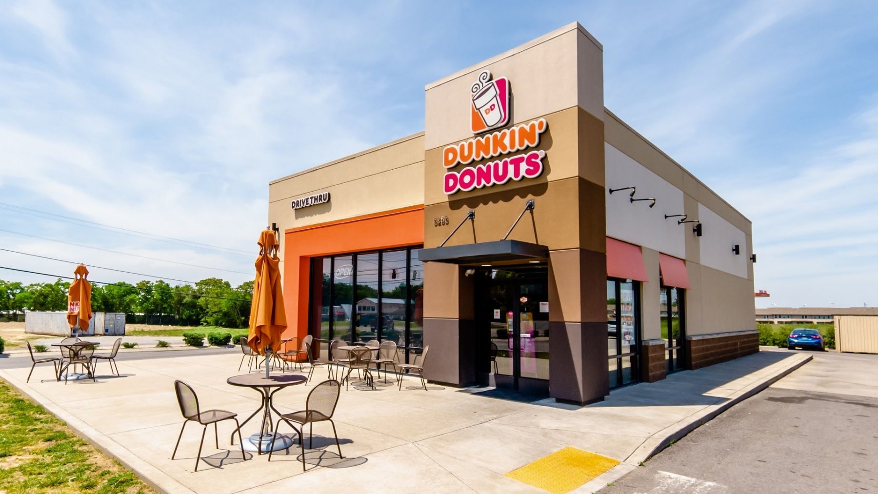 Dunkin' Donuts storefront, tan and orange building, outdoor seating area, blue sky.