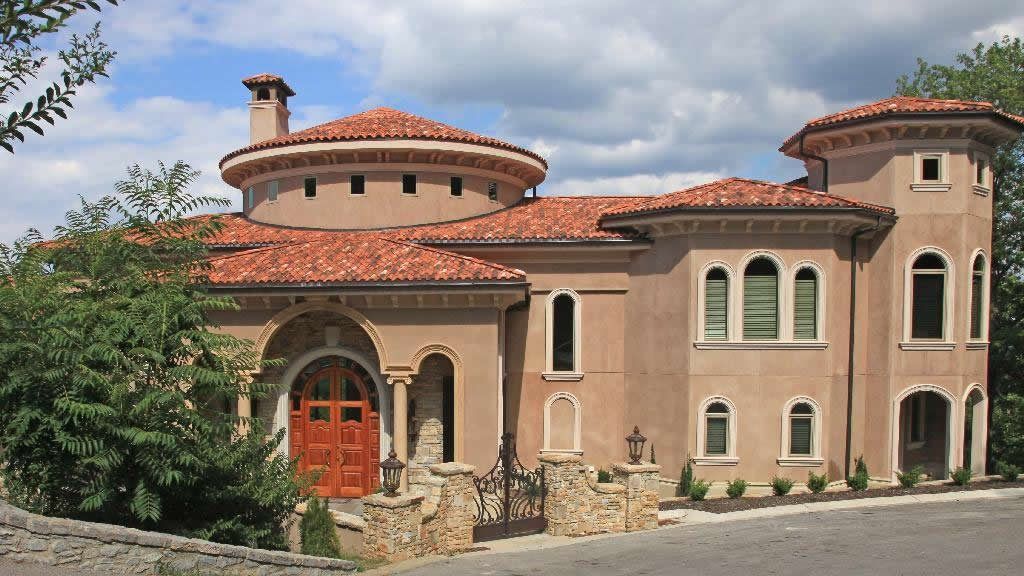 Beige stucco home with red tile roof, circular dome, and tower; surrounded by greenery and blue sky.