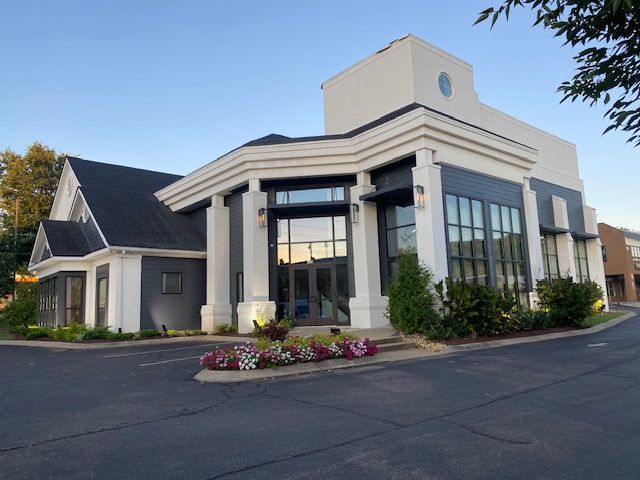 Modern building with glass front and white columns, surrounded by pavement and flowerbeds.