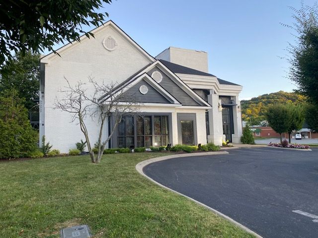 White building with dark-framed windows, surrounded by a curving asphalt drive and green lawn.