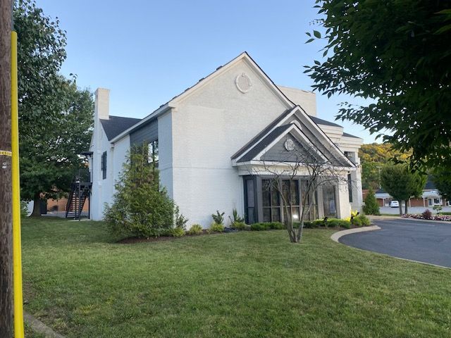 White building with dark roof, trees, and green lawn.