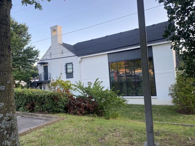 White building with black roof and large window, green bushes and trees in front.