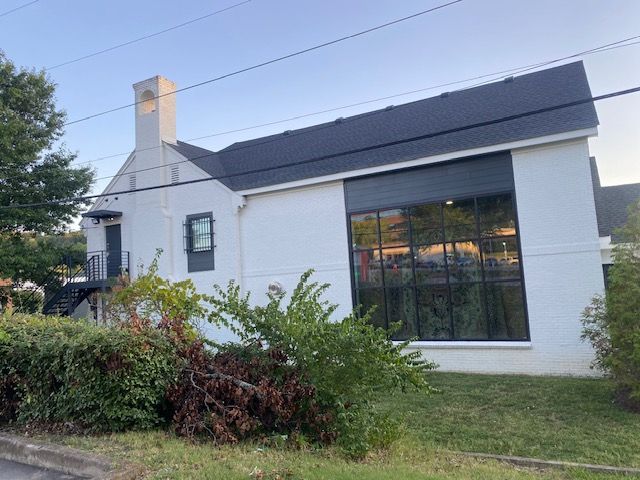 White building with black roof and large window. Green shrubs in front.