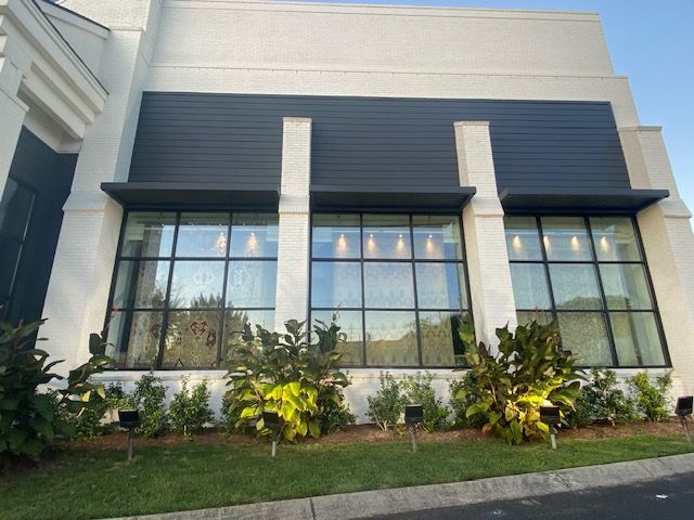 Exterior of a modern building with large windows and dark gray accents. Landscaping includes green bushes and grass.