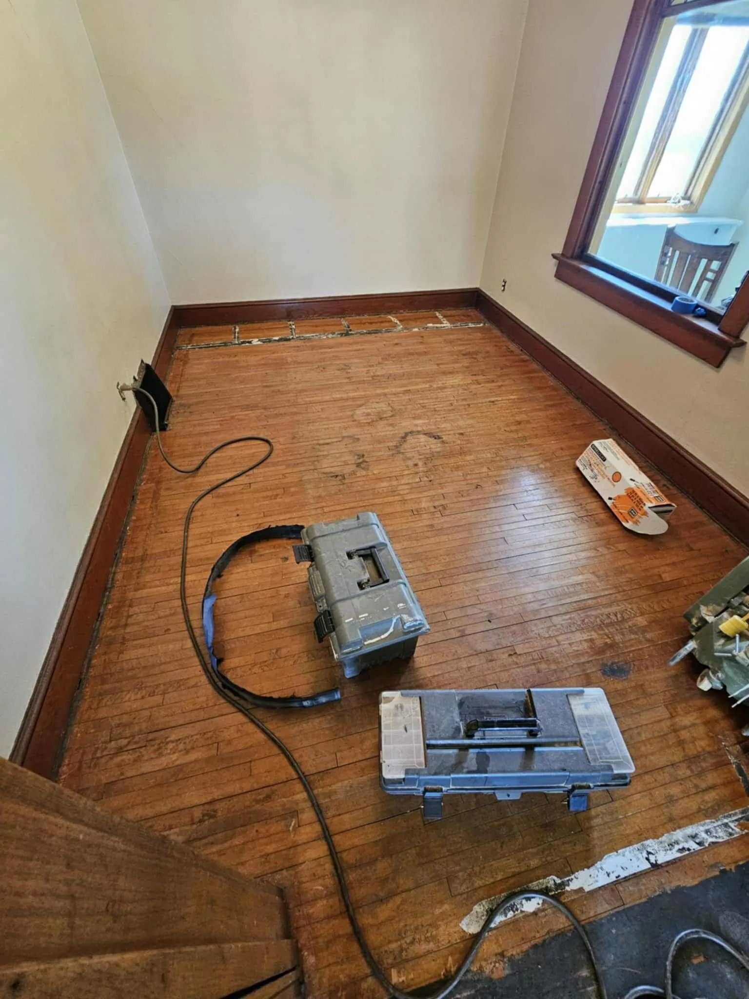 Empty room corner with exposed wood floor, tools, and cables near a window and wall outlet.
