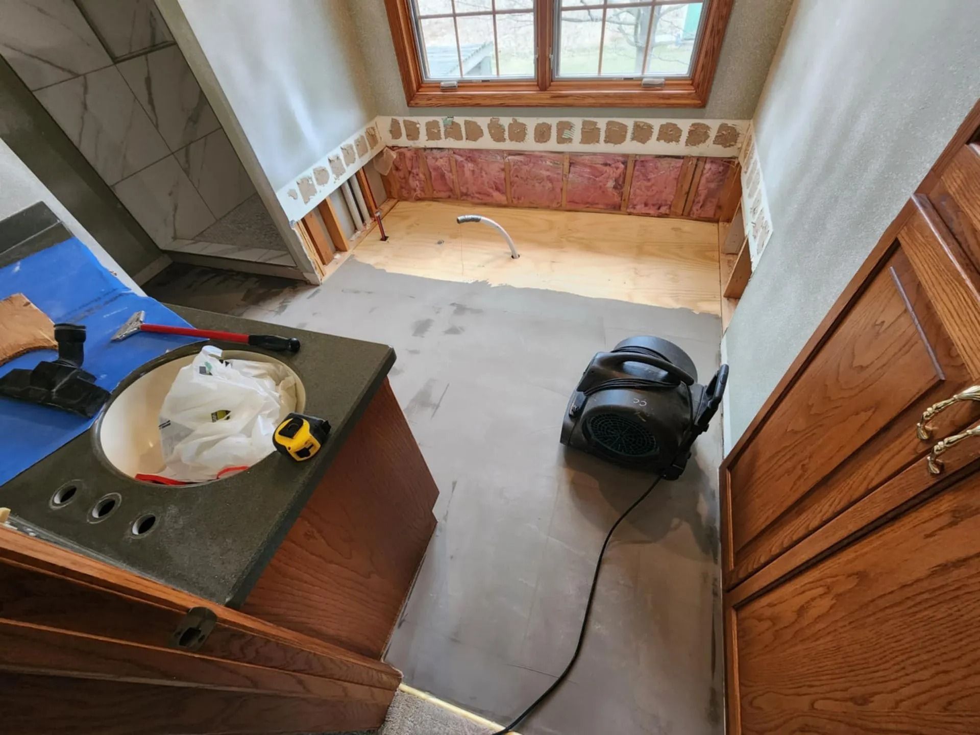 Small kitchen corner with sink, window, cabinets, a vacuum on the floor, and a plastic bag on the counter