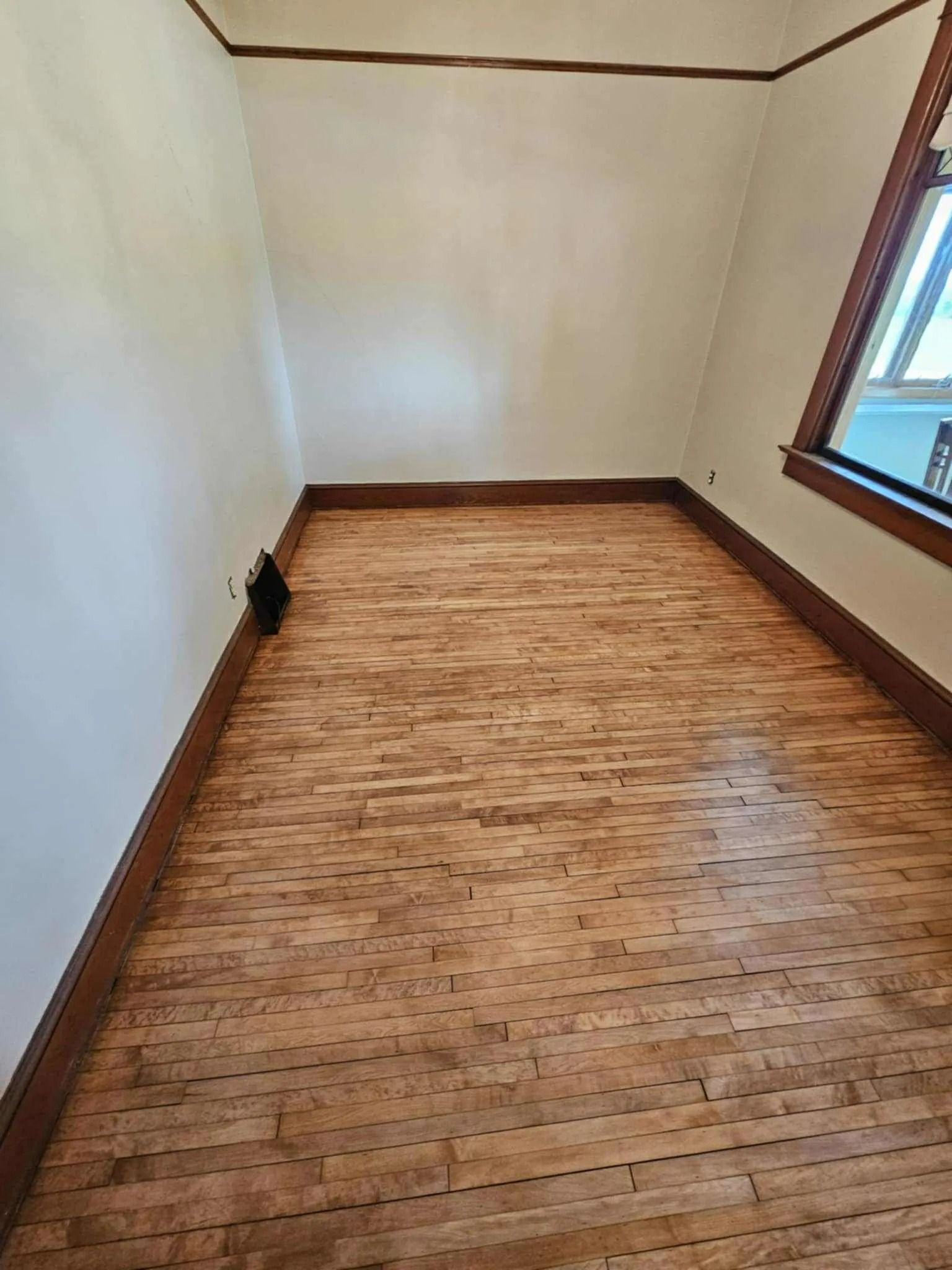 Empty small room with worn wood flooring, white walls, and a window on the right.