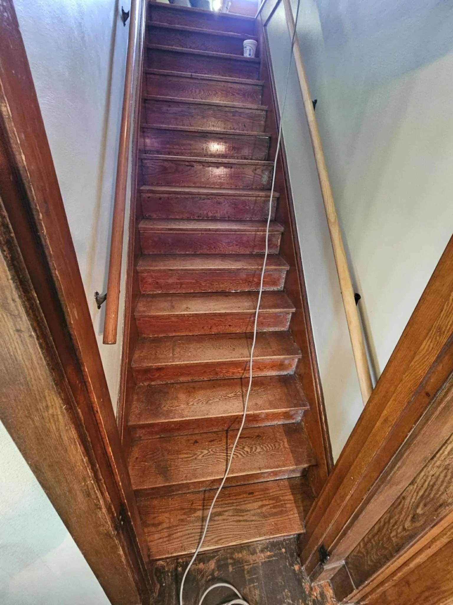 Narrow wooden staircase with a rope handrail, viewed from the bottom between white walls and wood trim