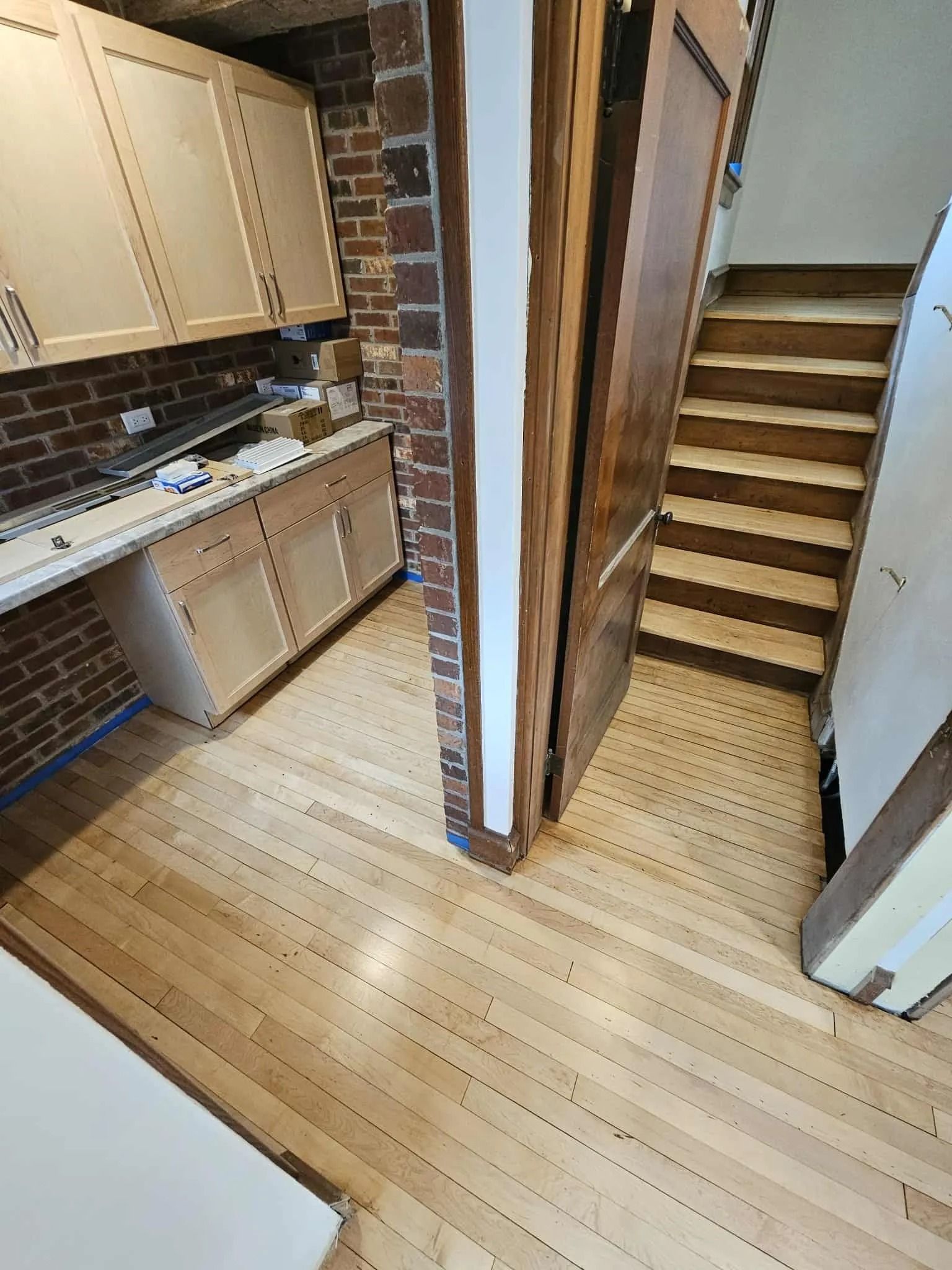 Small kitchen and staircase with wood cabinets, brick column, and light wood floor.