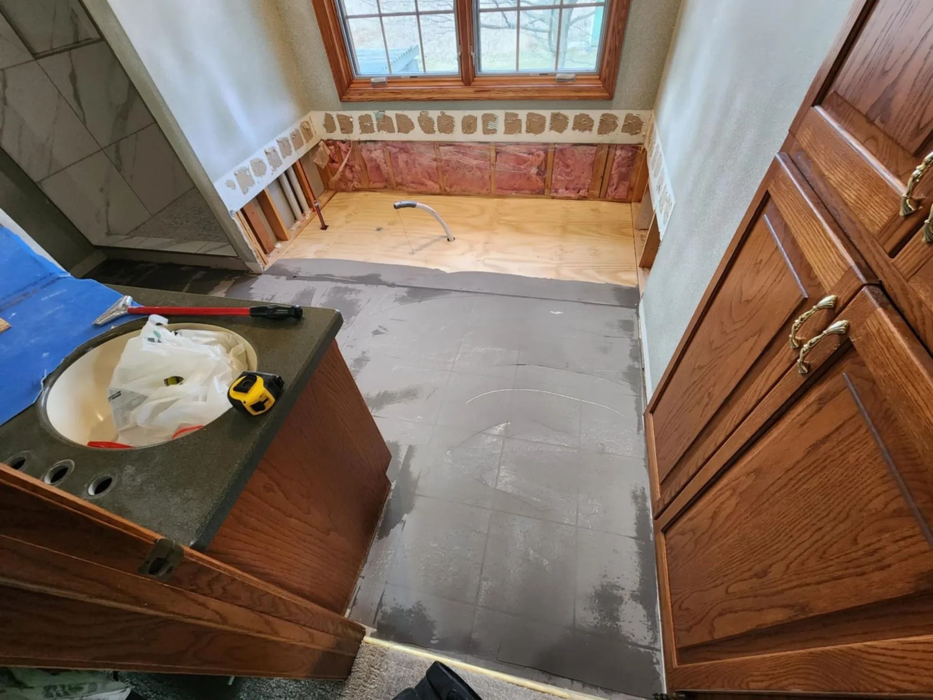 Partially renovated bathroom with gray floor patch, wood cabinets, and a sink under a window