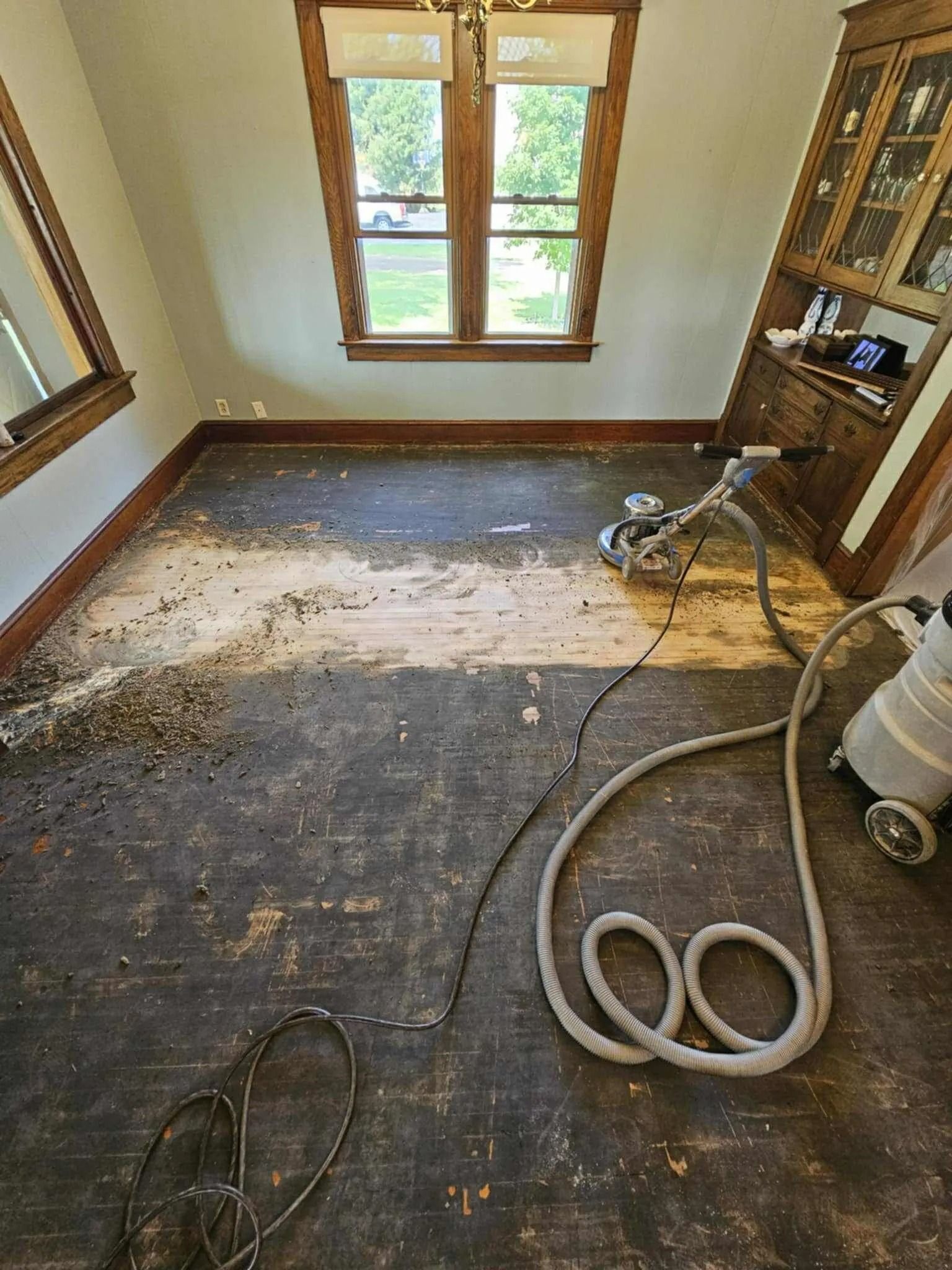 Dusty room with stripped carpet, a vacuum hose, and wood-framed windows along the back wall.