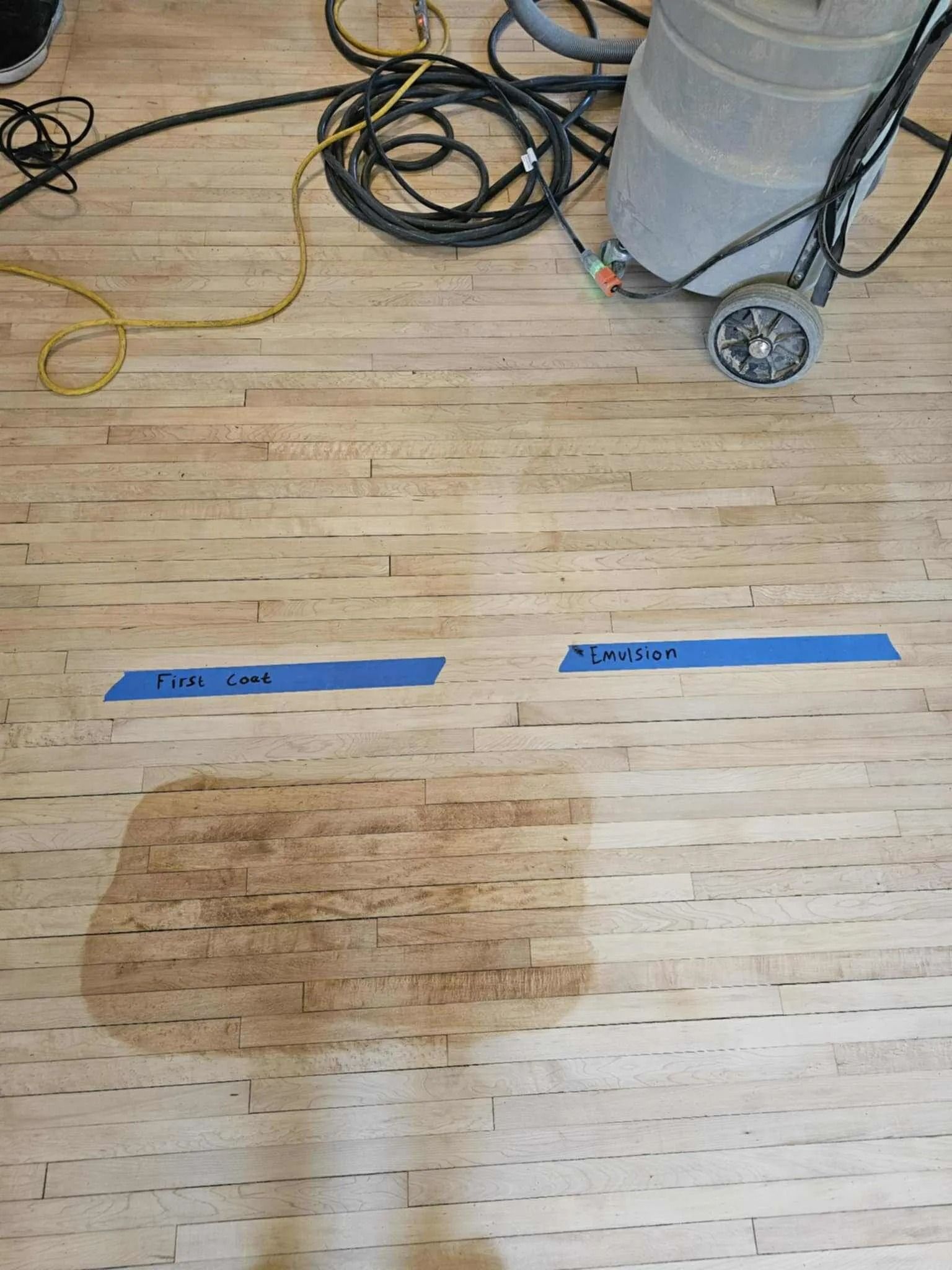 Wooden floor with two blue tape markers and a large worn patch near a wheeled machine base.