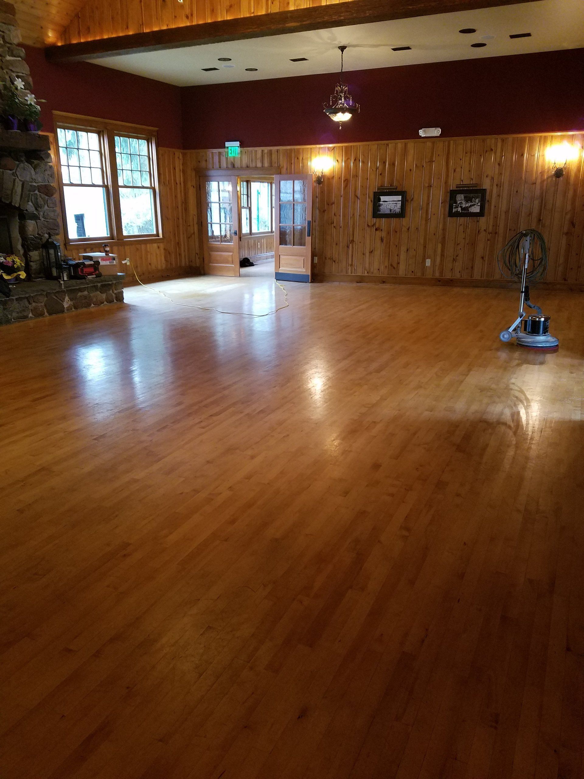 Empty wooden dance hall with polished floor, windows, and a ceiling fan near the back wall.