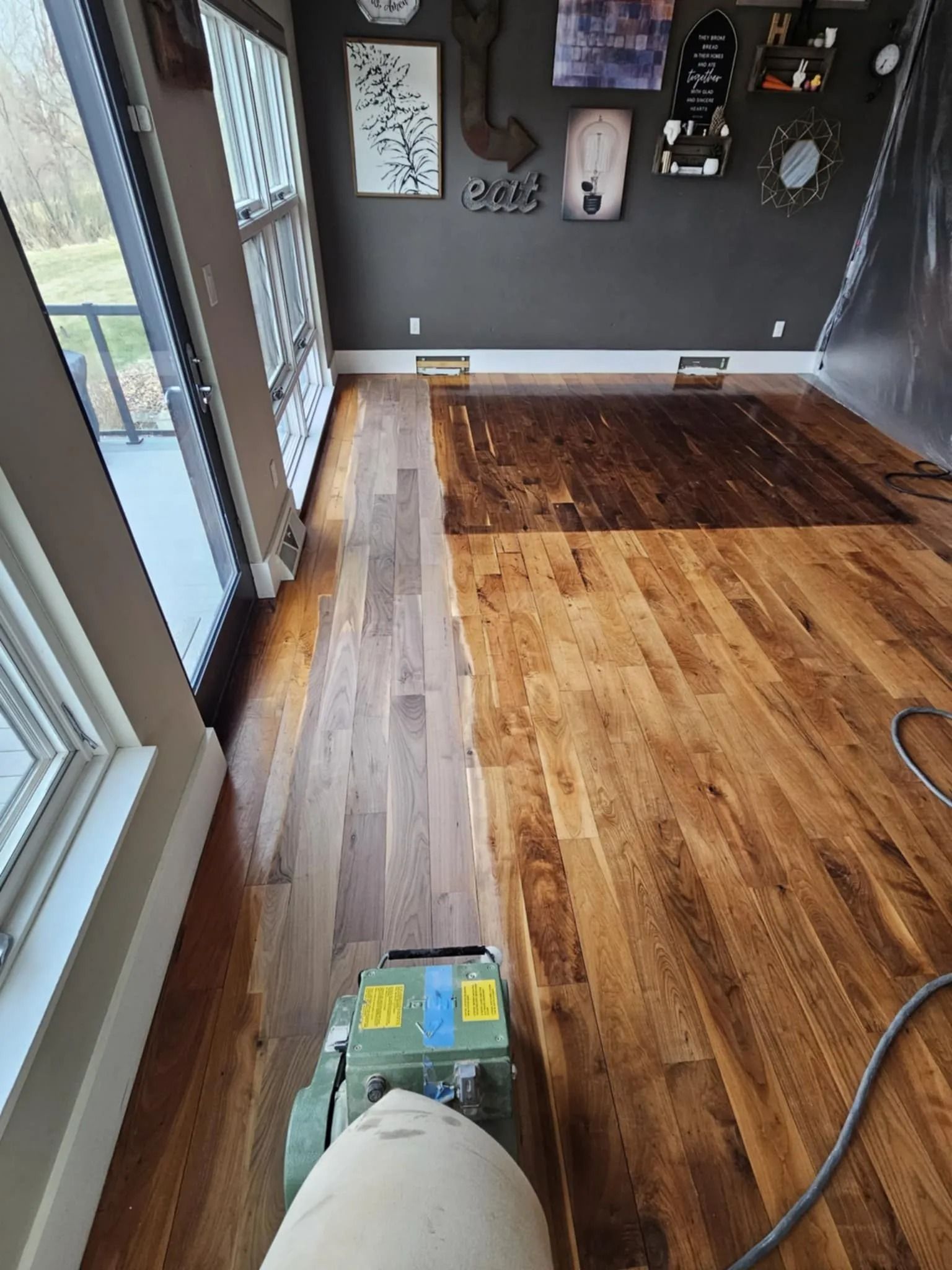 Hallway with hardwood floor being sanded, showing a floor sander and dust hose near glass doors.