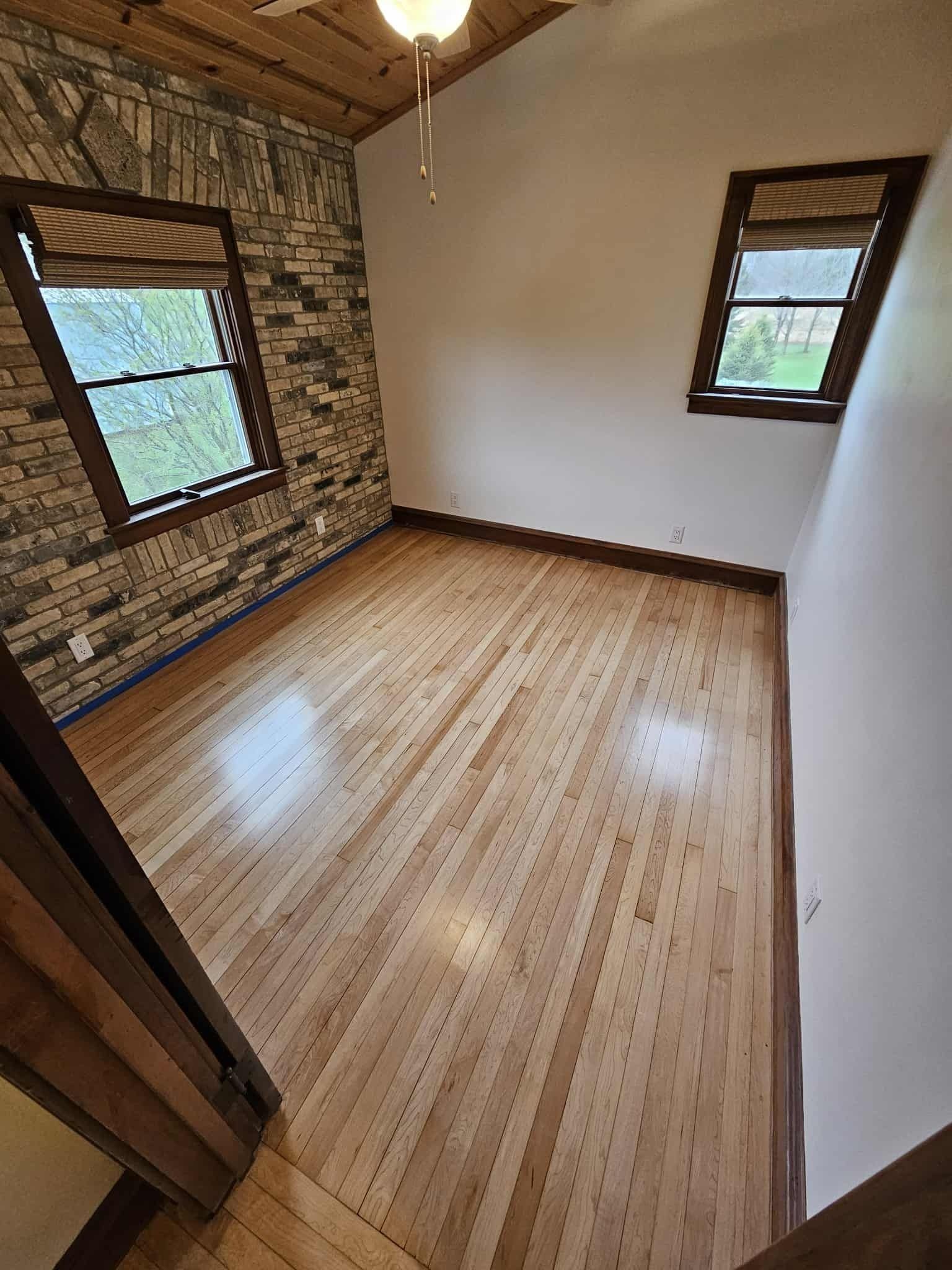 Empty room with wood plank floor, exposed brick wall, white wall, and two windows.