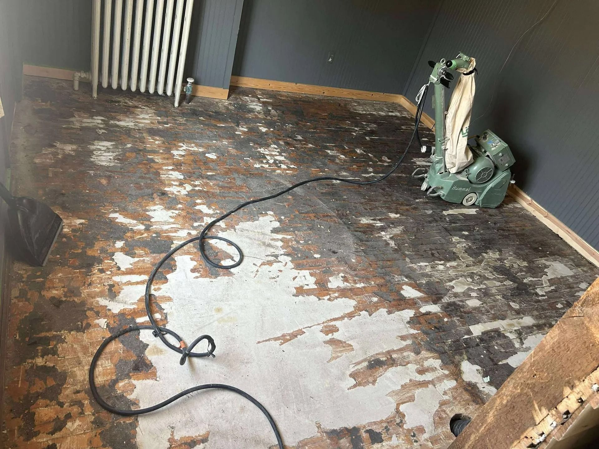 Stripped floor in a room with a sander and power cord, with patches of old flooring removed.