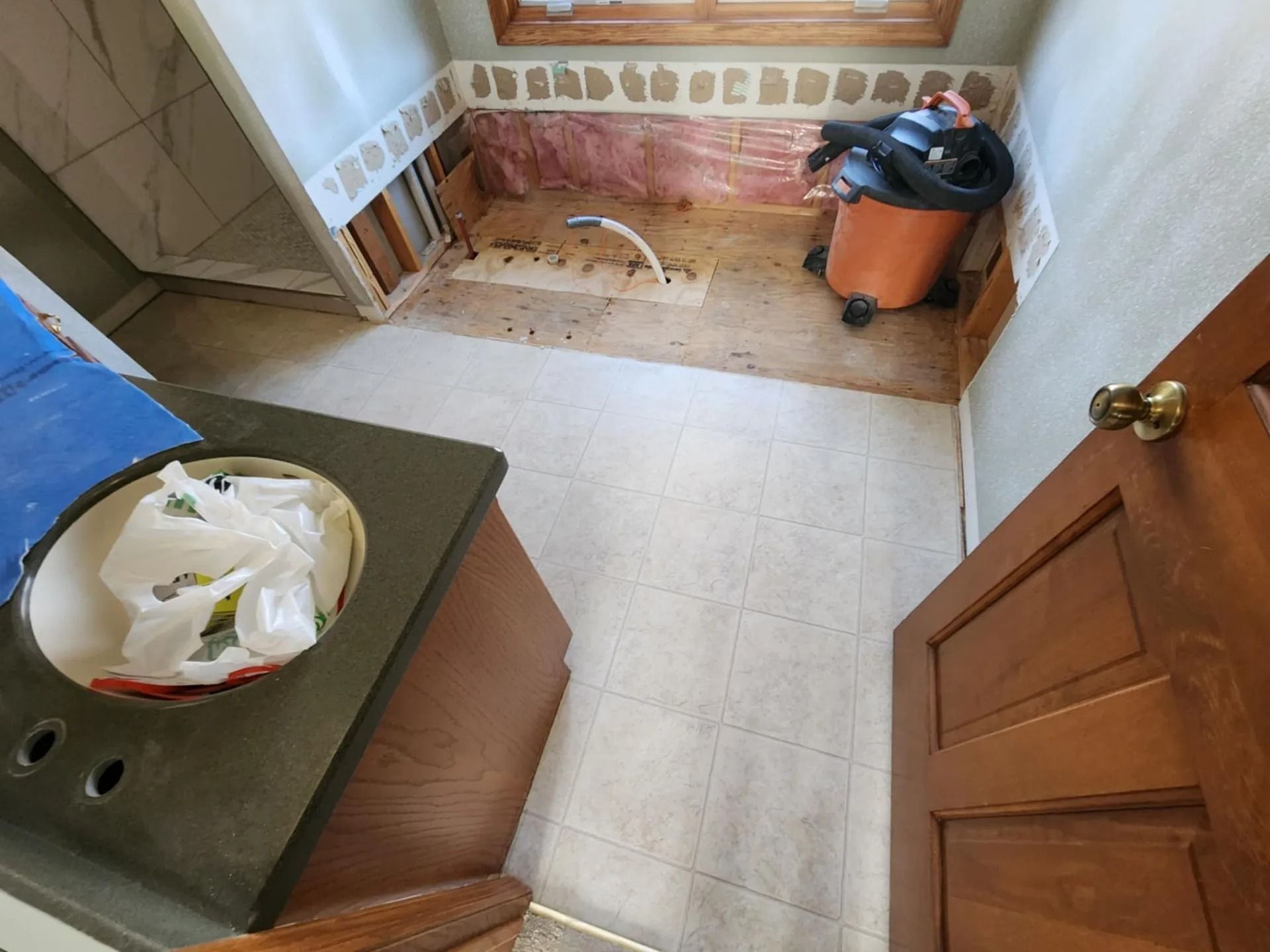 Small room under renovation with tiled floor, exposed wall, tools, and an orange shop vacuum near the back.