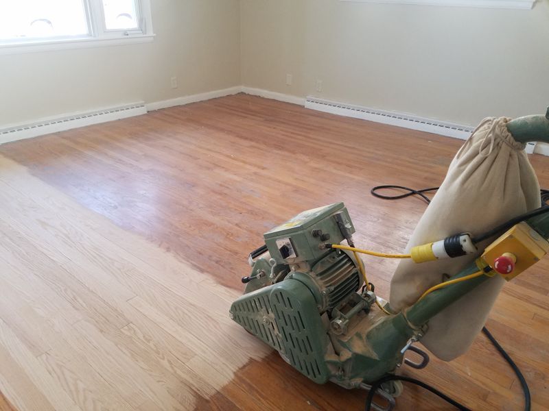 Floor sander in a bare room, refinishing a wood floor with a fresh sanded section visible.