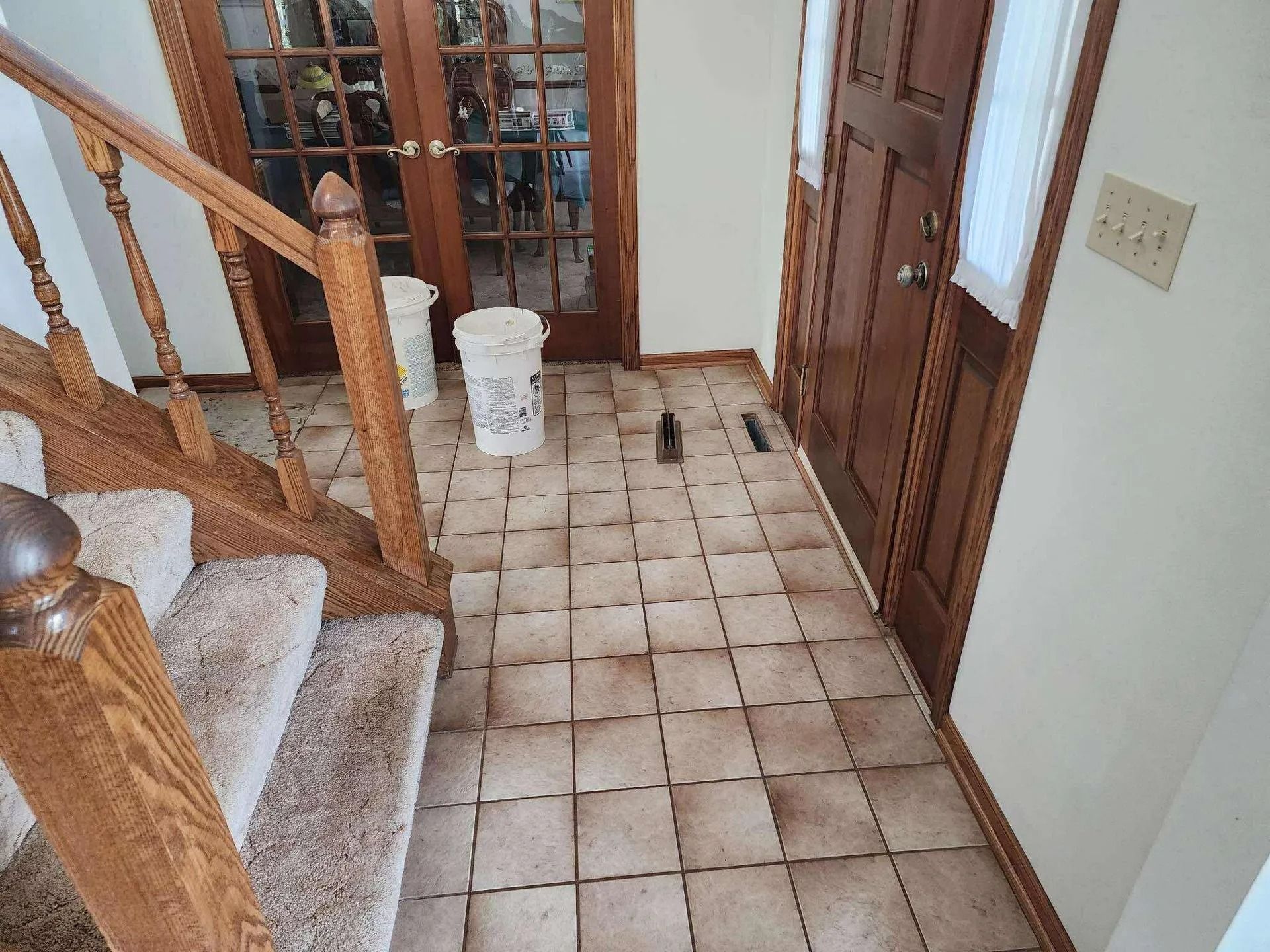 Stairway landing with beige tile floor, wooden banister, and a white bucket near the French doors.