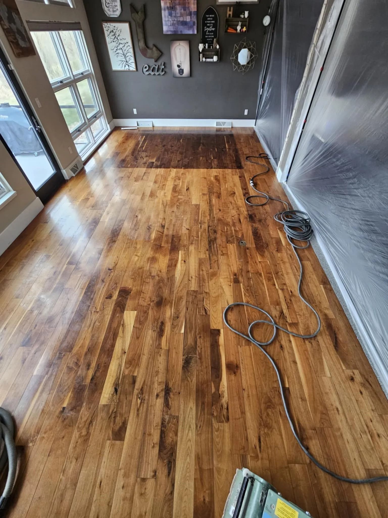 Empty hardwood room with scattered cords, gray walls, and sunlight from glass doors.