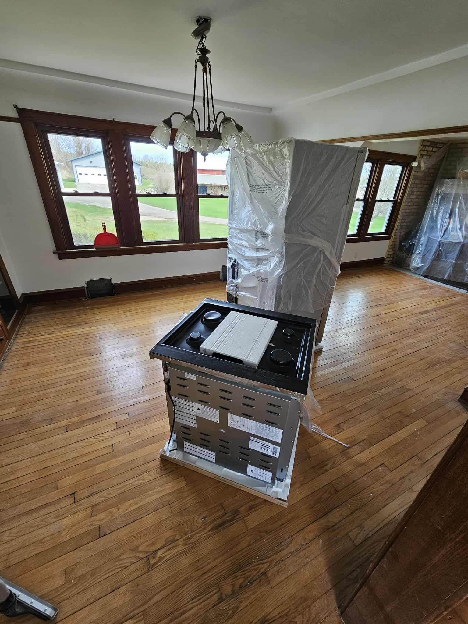 Kitchen with a large wrapped appliance on a box in an empty room with wooden floors and windows