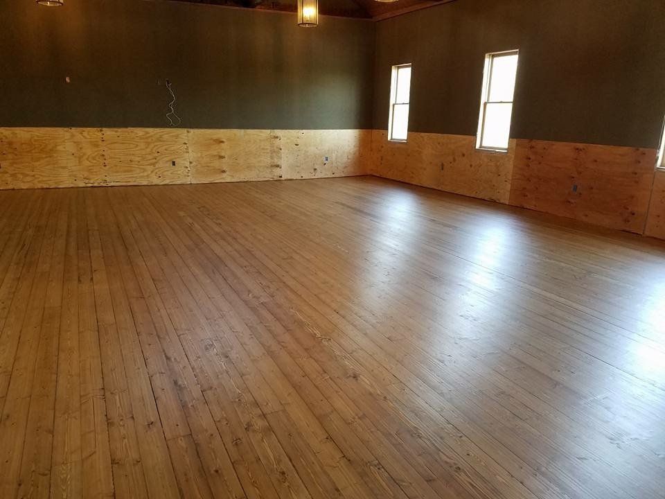 Empty wooden room with polished floor, paneled walls, and two tall windows letting in light