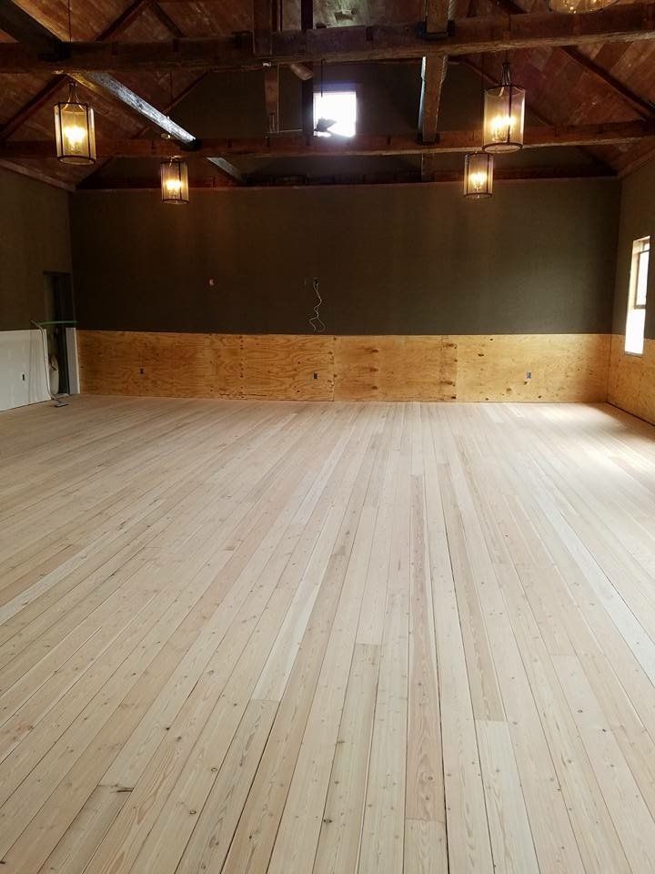 Empty wooden hall with exposed beams and hanging lights, viewed from the back toward a dark stage wall.