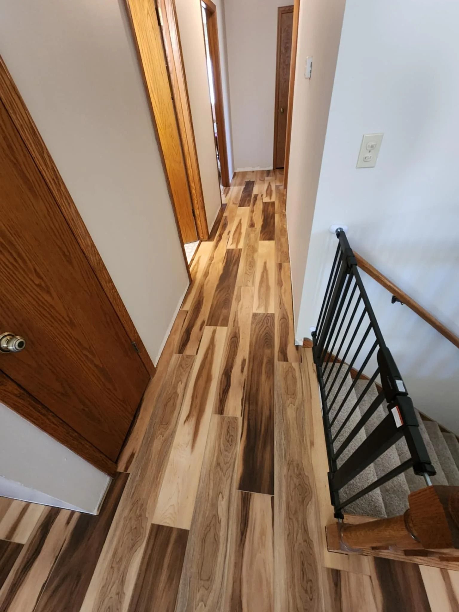 Narrow hallway with wood floors, white walls, and a black stair railing on the right.