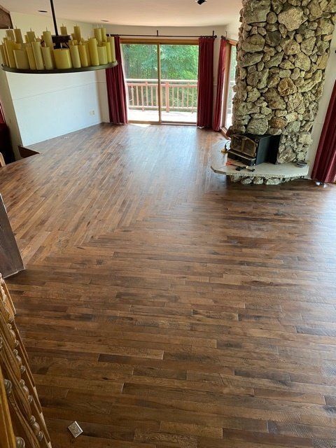 Empty living room with hardwood floors, stone fireplace, and balcony doors overlooking trees