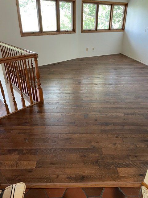 Empty hardwood-floored room with white walls, windows, and a wooden staircase railing.