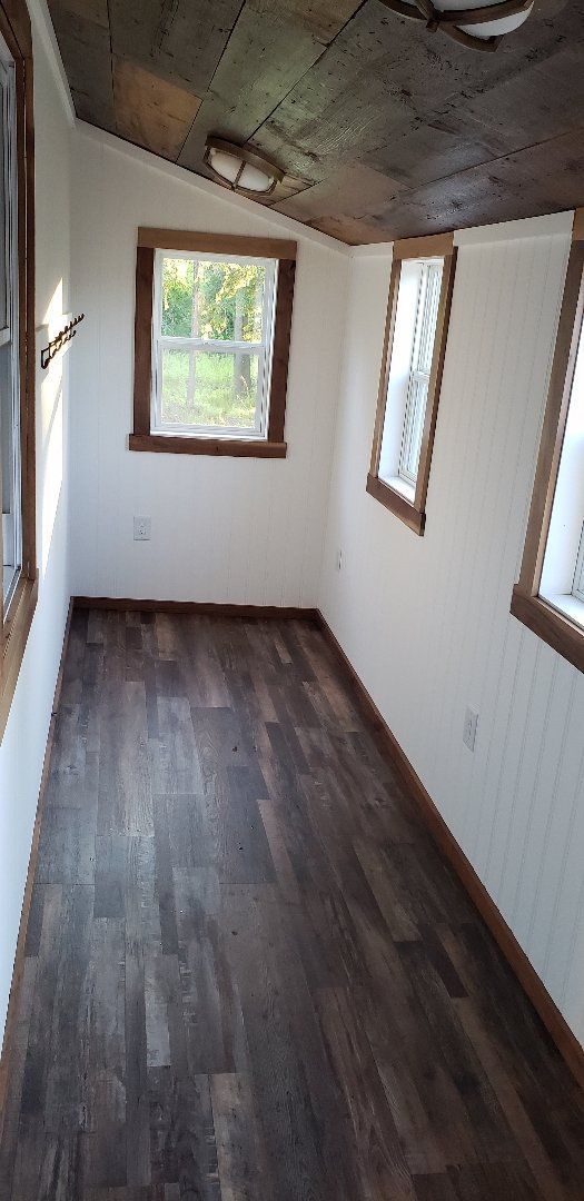 Narrow empty room with dark wood floor, white walls, and two small windows, one showing greenery outside