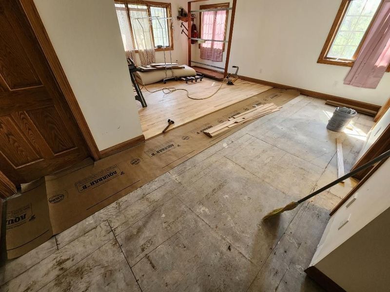 Interior room under renovation with bare floorboards, scattered tools, and windows letting in daylight