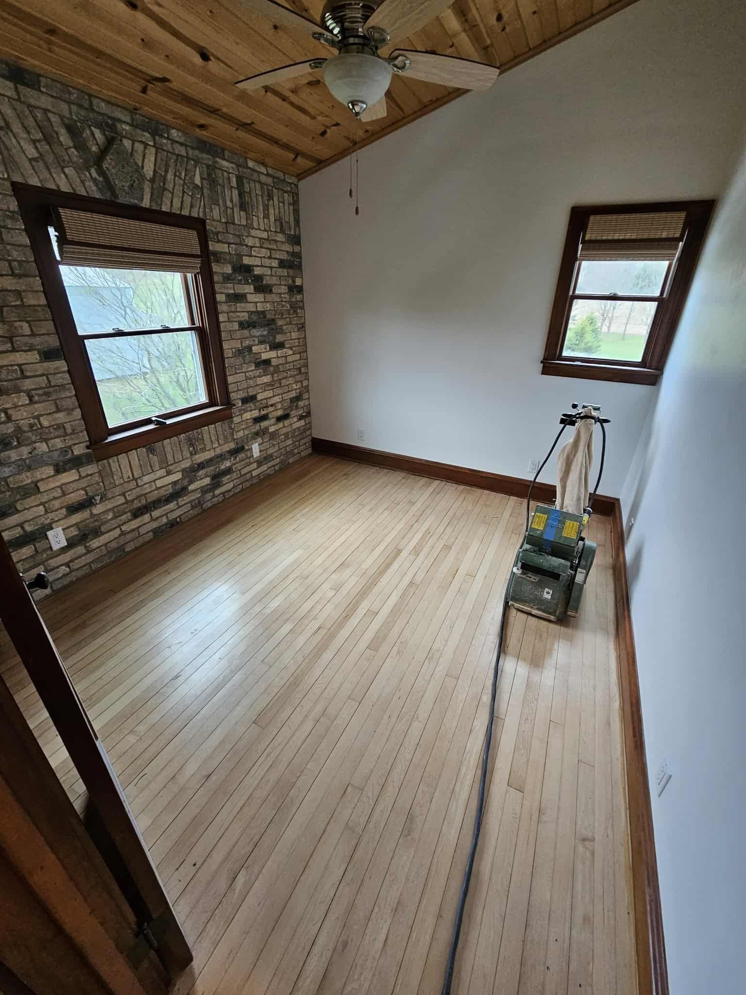 Empty room with wood floors, exposed wood ceiling, brick wall, and a paint sprayer on the floor.
