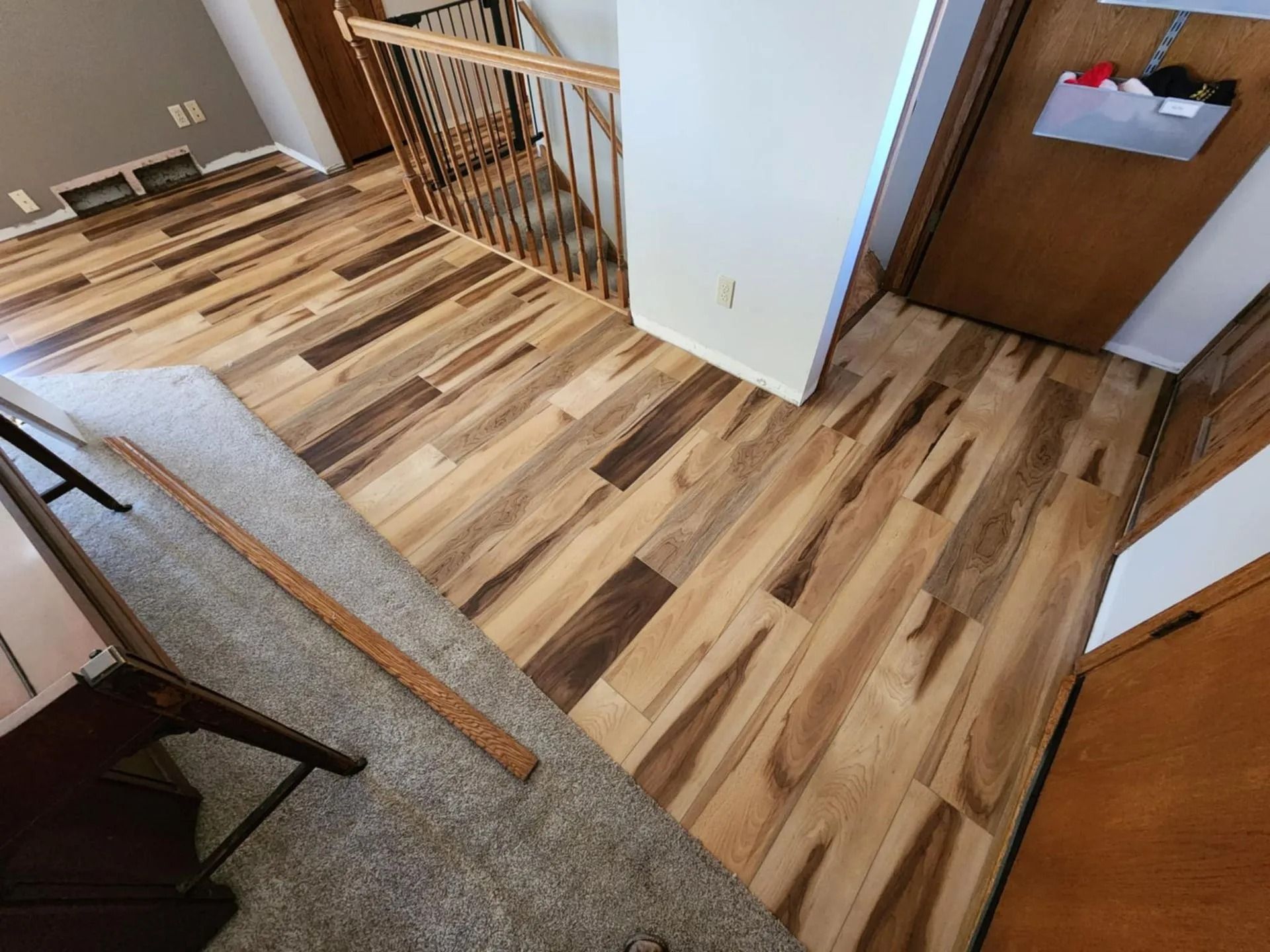 Interior hallway with wood-patterned flooring, carpeted stairs, and a doorway with papers on the wall.