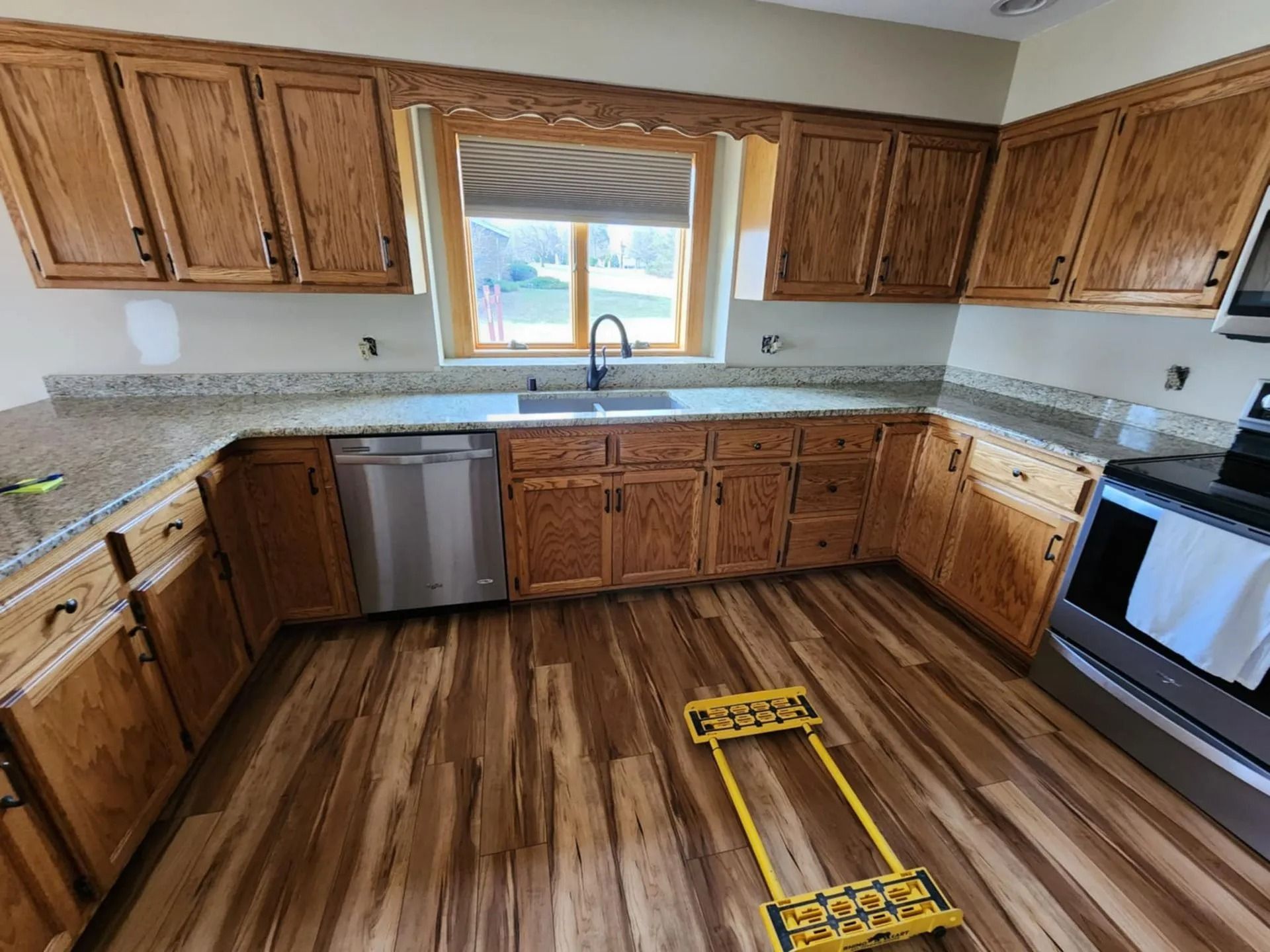 Empty kitchen with wood cabinets, granite counters, stainless appliances, and a window over the sink.