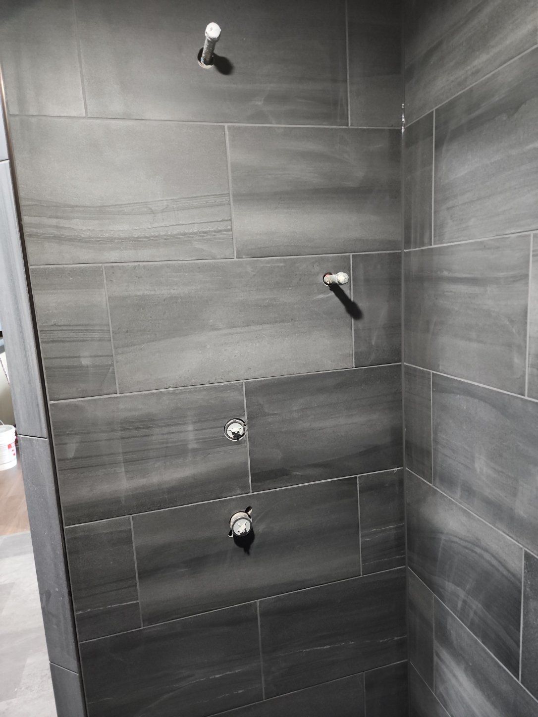 Gray tiled shower wall with three shower fixtures and a showerhead, viewed from inside a bathroom.